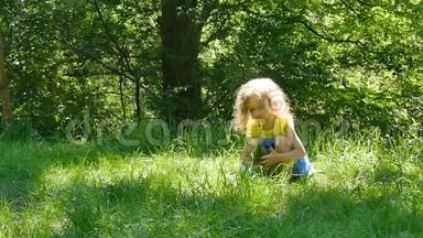 小可爱的金发女孩穿着明亮的黄色衬衫和蓝色的裙子<strong>坐在公园里</strong>的绿草上，把它捡起来。